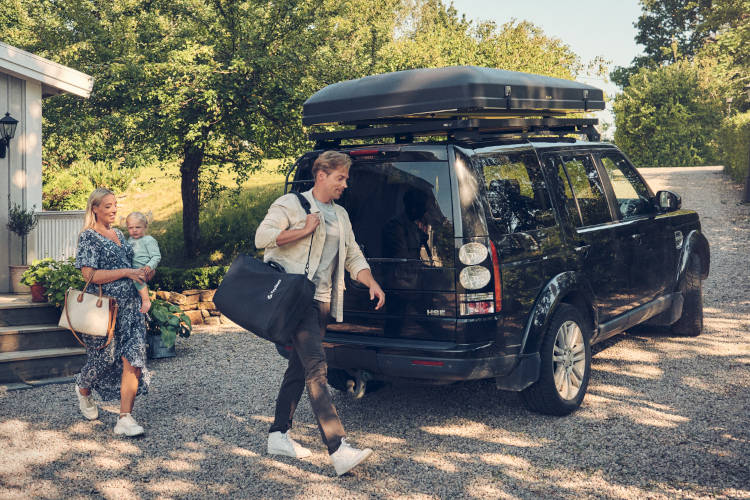 Man is carrying a TinySeats Two in a shoulder bag. Behind him is his wife carrying their baby. They are about to install their TinySeats Two in their Land Rover. It is Swedish summer